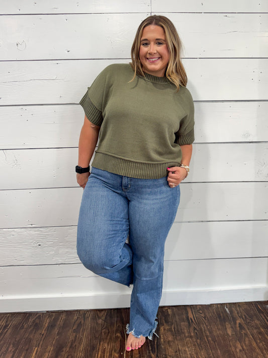 A woman modeling an olive green short-sleeve mock neck knit sweater with ribbed cuffs and hem, paired with blue jeans, standing against a white wood-paneled wall.