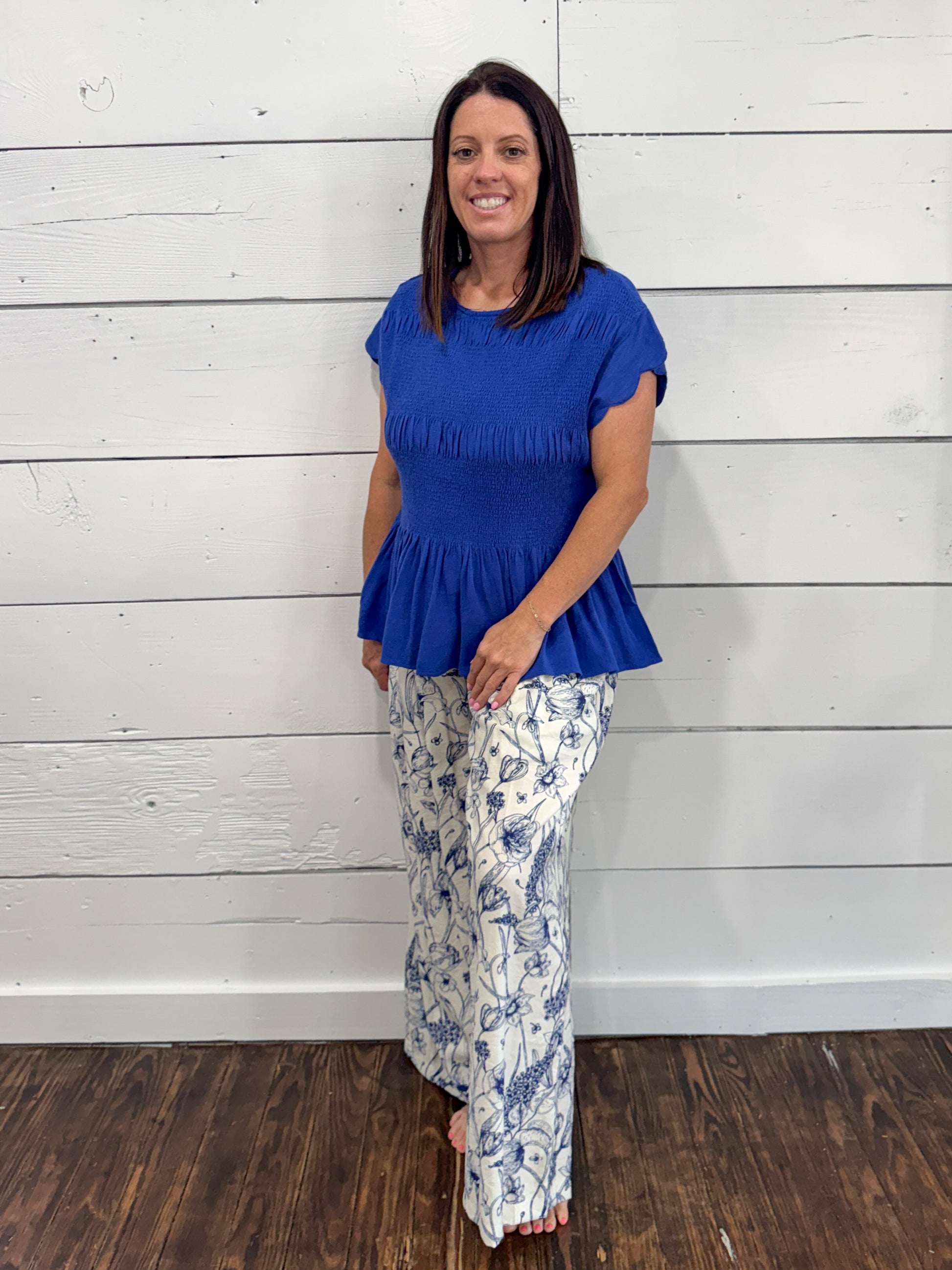 A woman modeling a royal blue short-sleeve peplum top and white pants with a blue floral toile pattern, standing against a white wood-paneled wall.