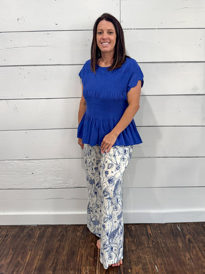 A woman modeling a royal blue short-sleeve peplum top and white pants with a blue floral toile pattern, standing against a white wood-paneled wall.