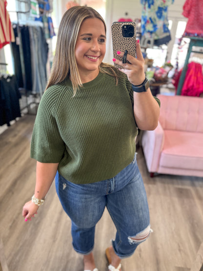 A woman takes a mirror selfie in a clothing boutique wearing an olive green short-sleeve ribbed knit sweater and blue jeans.
