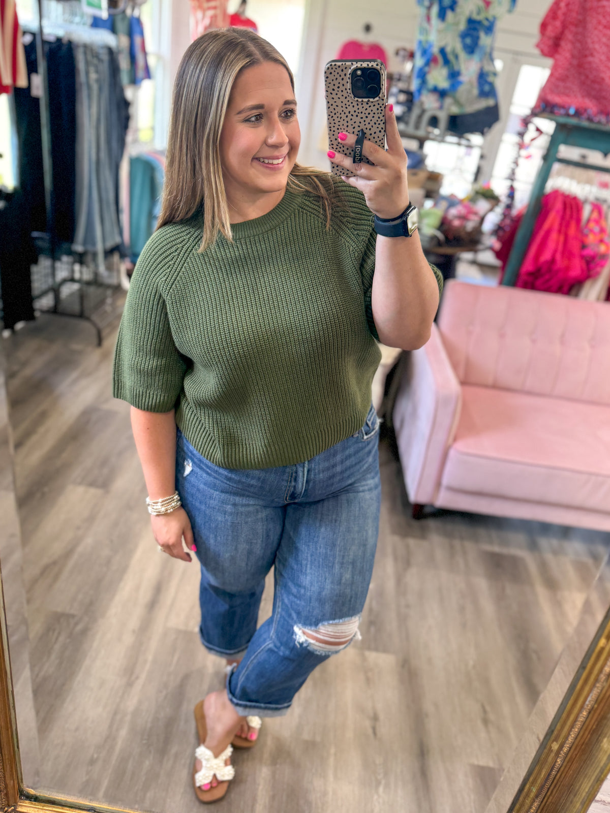 A woman takes a mirror selfie in a clothing boutique wearing a short-sleeve ribbed olive green top, distressed blue jeans, and white slide sandals.