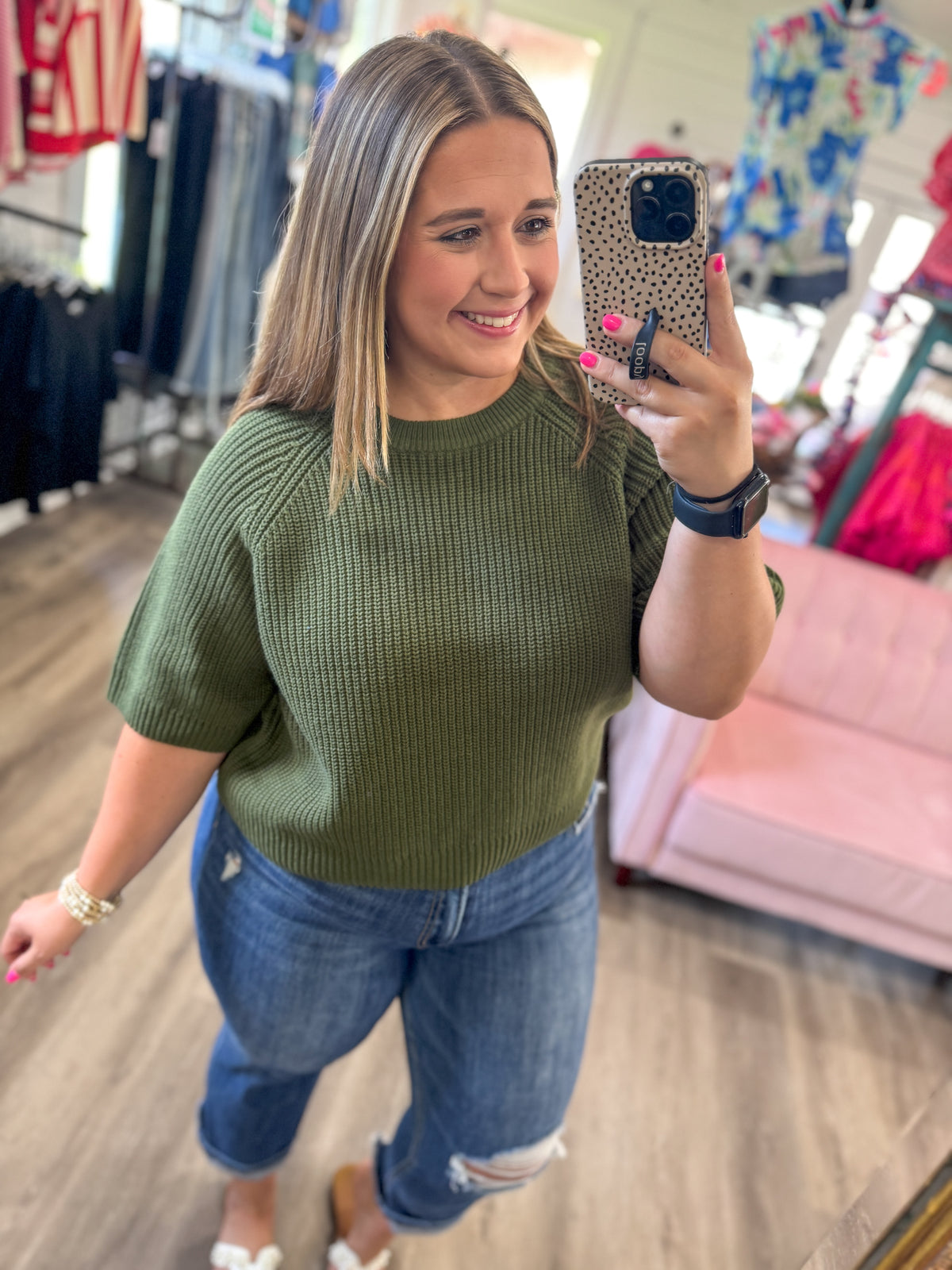 A woman takes a mirror selfie in a clothing store, wearing a ribbed short-sleeve crewneck sweater in olive green and distressed blue jeans.