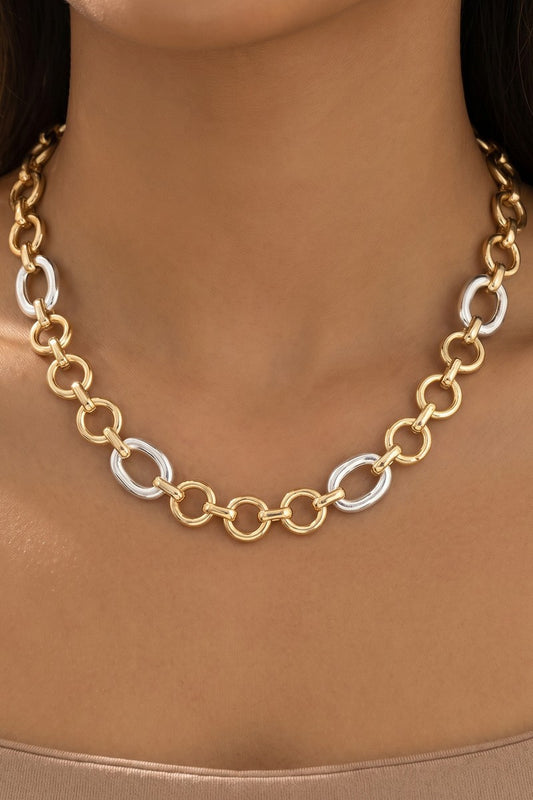 A close-up of a model wearing a gold and silver link necklace. The necklace features alternating large gold oval links and smaller silver oval links, shown against the model's neck.
