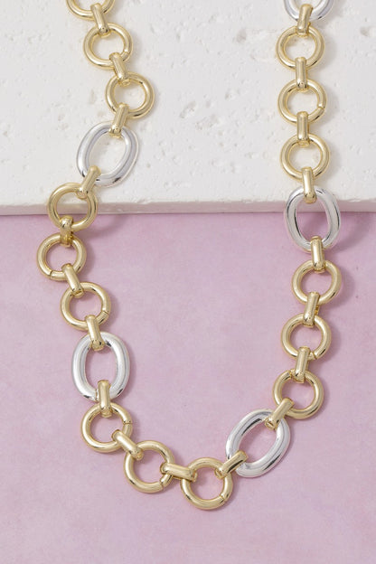 A two-tone chain necklace featuring alternating gold and silver oval links, displayed on a split white and light pink background.