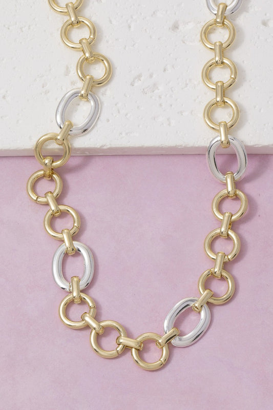 A two-tone chain necklace featuring alternating gold and silver oval links, displayed on a split white and light pink background.