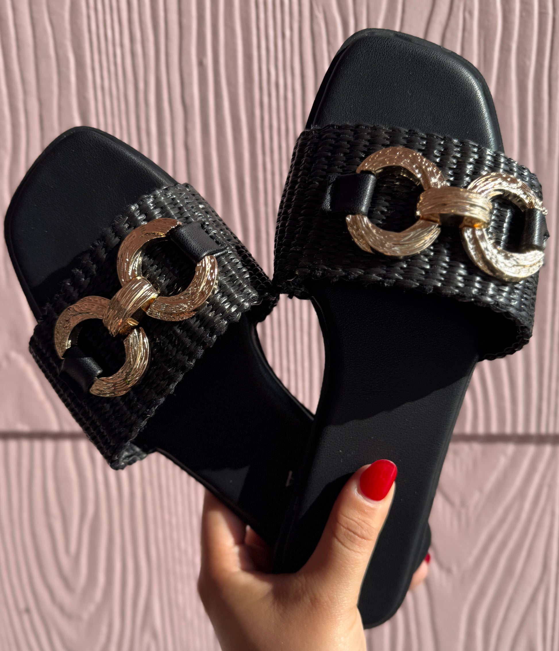 Black slides with gold chain details held by a hand against a pink background