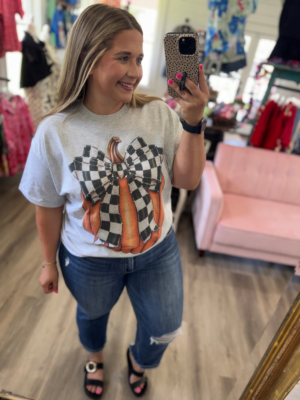 Woman taking a mirror selfie wearing a graphic t-shirt with pumpkins and checkered pattern in a store.