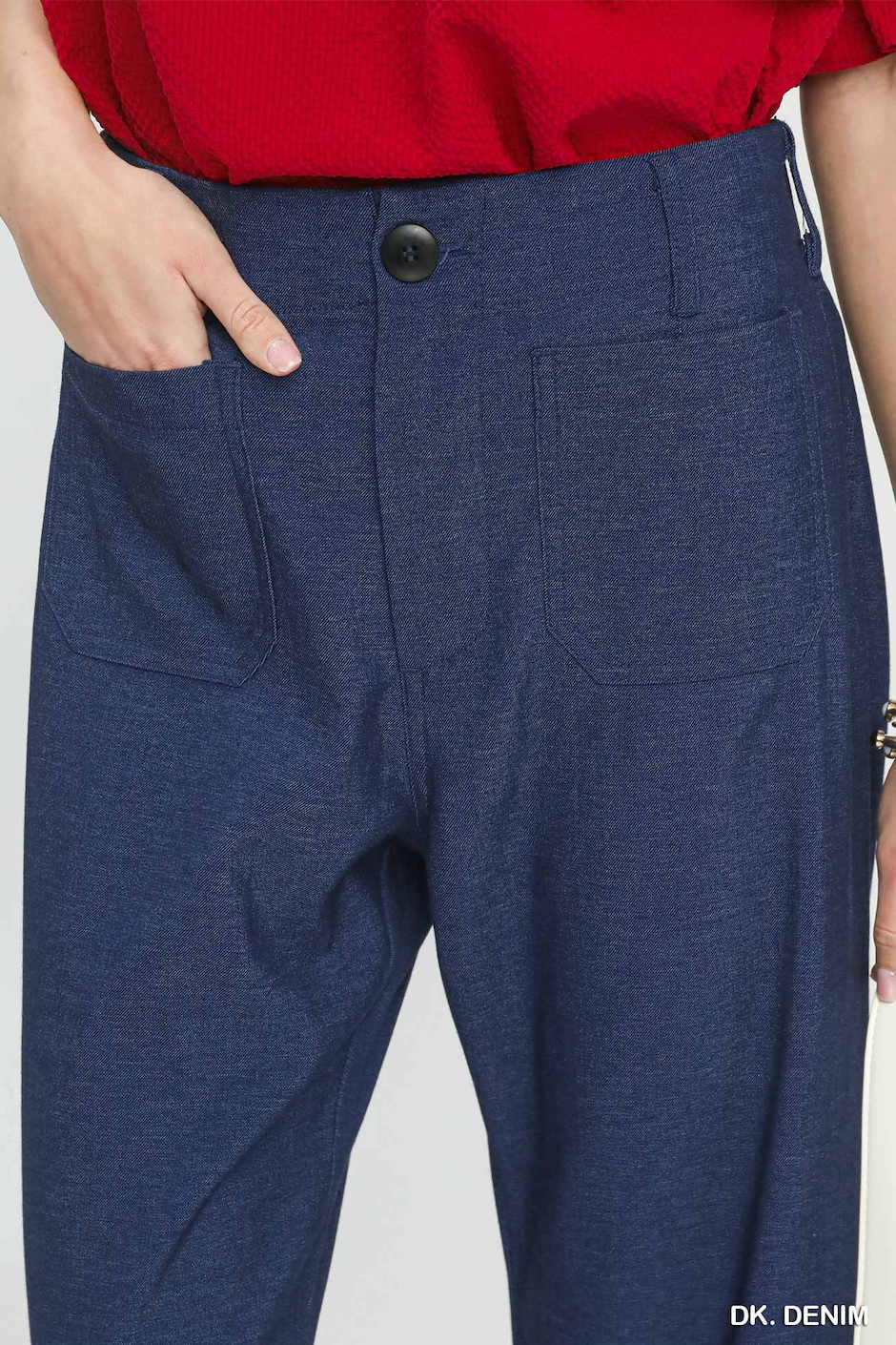 Close-up of a person wearing dark blue heathered trousers with a red top, showing the front waistband, button, side pockets, and hand in pocket against a light gray background.