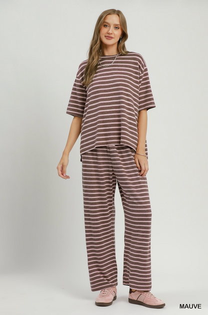 A woman modeling a matching two-piece lounge set consisting of an oversized short-sleeve t-shirt and wide-leg pants, both in a brown and white horizontal stripe pattern. She is wearing pink slip-on shoes against a plain light gray studio background.