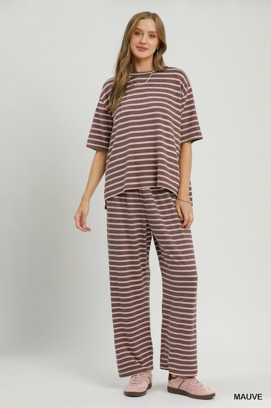 A woman modeling a matching two-piece lounge set consisting of an oversized short-sleeve t-shirt and wide-leg pants, both in a brown and white horizontal stripe pattern. She is wearing pink slip-on shoes against a plain light gray studio background.