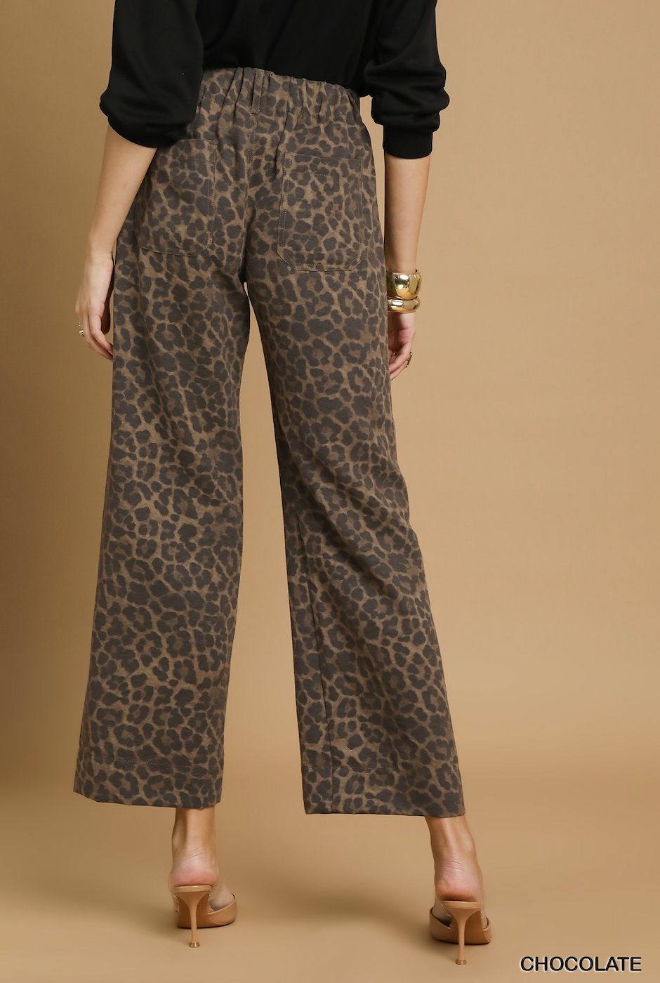 Rear view of a model wearing wide-leg cropped pants with a brown and beige leopard print, paired with a black long-sleeve top and tan high heels, against a solid tan studio background.