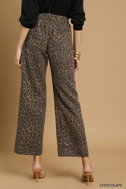 Rear view of a model wearing wide-leg cropped pants with a brown and beige leopard print, paired with a black long-sleeve top and tan high heels, against a solid tan studio background.