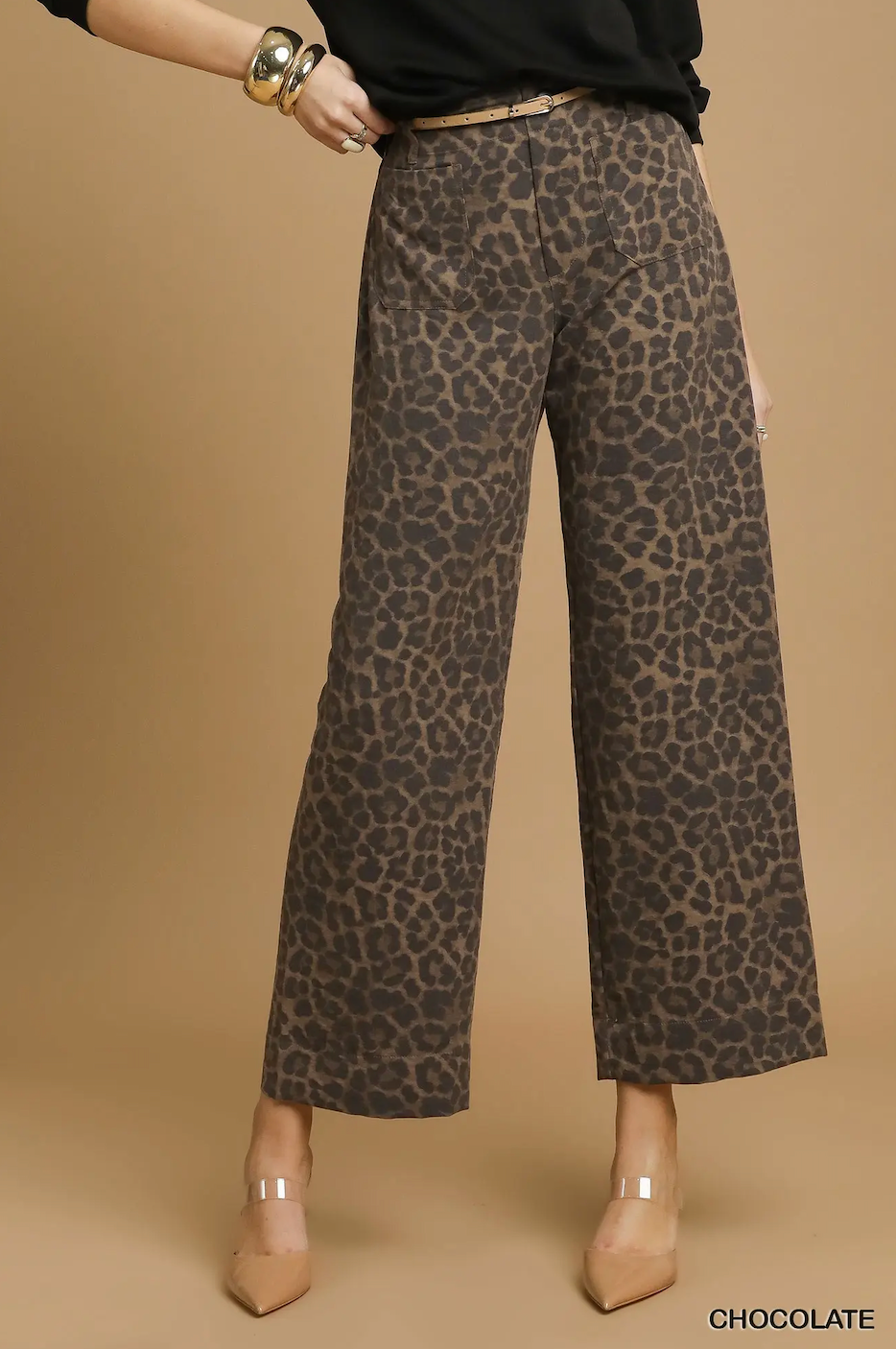 A model wearing high-waisted, wide-leg cropped pants in a brown and beige leopard print pattern, styled with a black top, beige heels, and gold jewelry against a solid tan background.