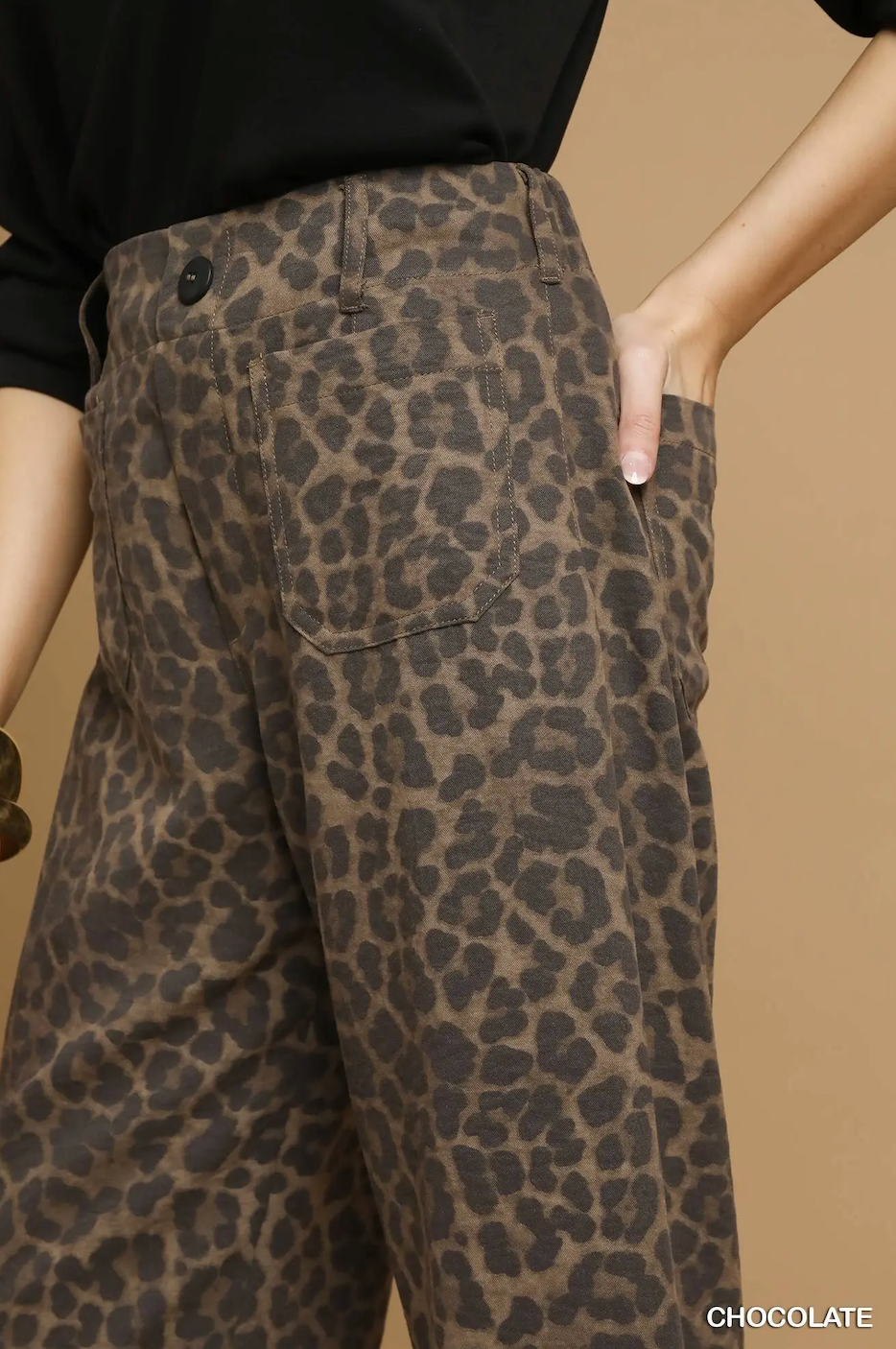 Close-up of a person wearing wide-leg trousers with a brown and black leopard print pattern, paired with a black top against a warm beige studio background.