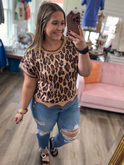A woman takes a mirror selfie in a boutique wearing a beige short-sleeve top with a brown leopard print pattern, paired with distressed blue jeans.