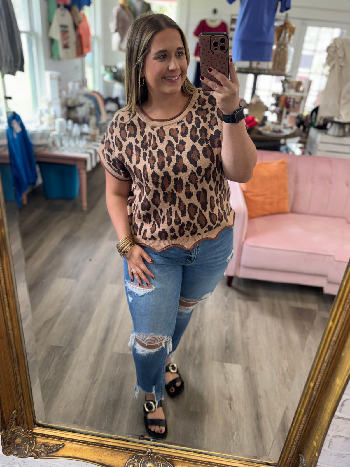 A woman takes a mirror selfie in a boutique, wearing a beige short-sleeve top with a brown leopard print pattern and distressed blue jeans.