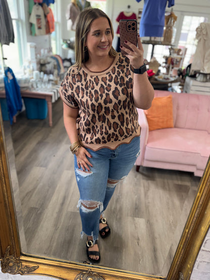A woman takes a mirror selfie in a boutique, wearing a beige short-sleeve top with a brown leopard print pattern and distressed blue jeans.