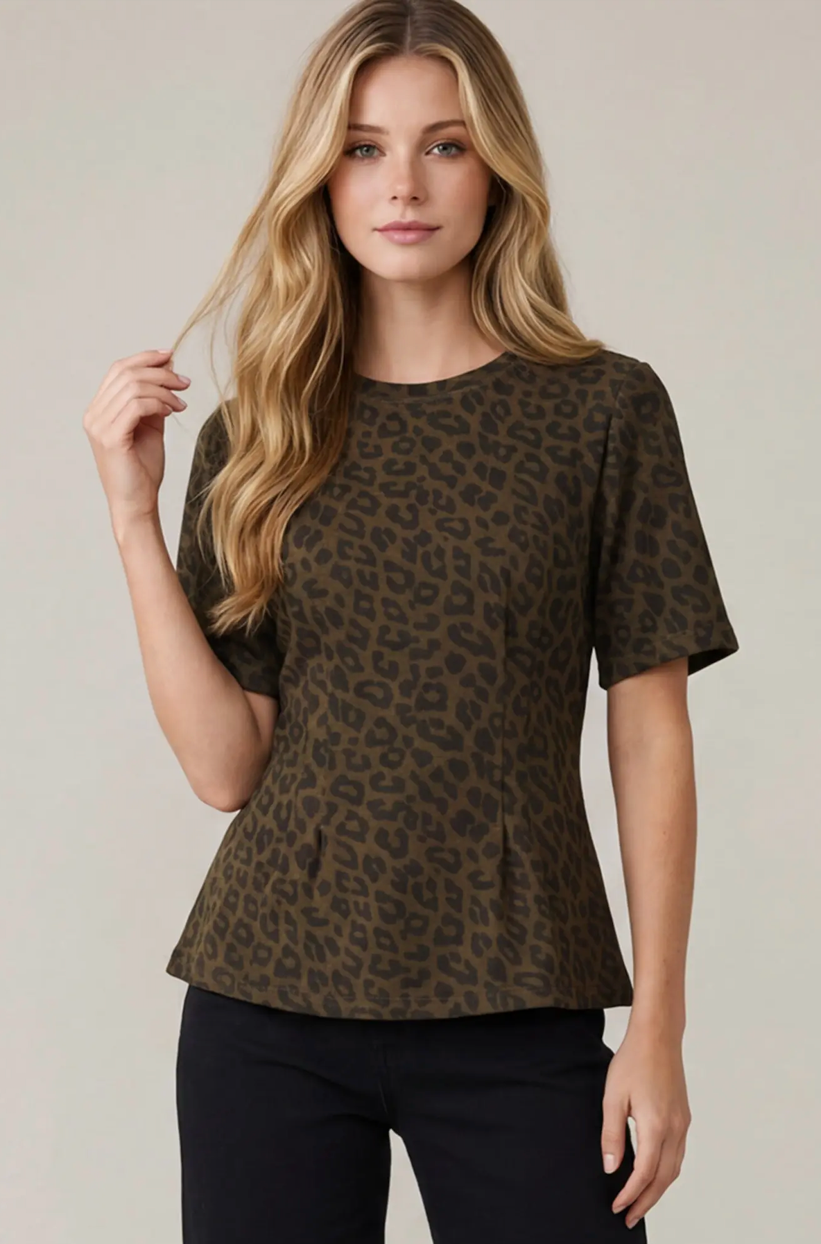 A woman modeling a short-sleeve crewneck top with an olive brown and black leopard print pattern, paired with black pants, against a solid light beige background.