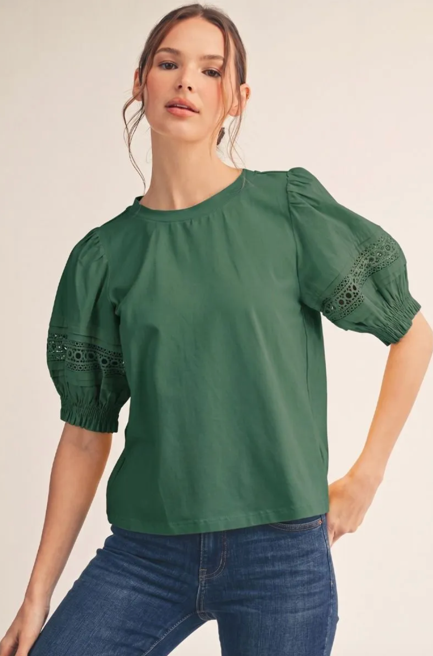 A woman wearing a green short-sleeve blouse with puff sleeves and eyelet lace detailing near the cuffs, paired with blue jeans, against a plain light beige studio background.