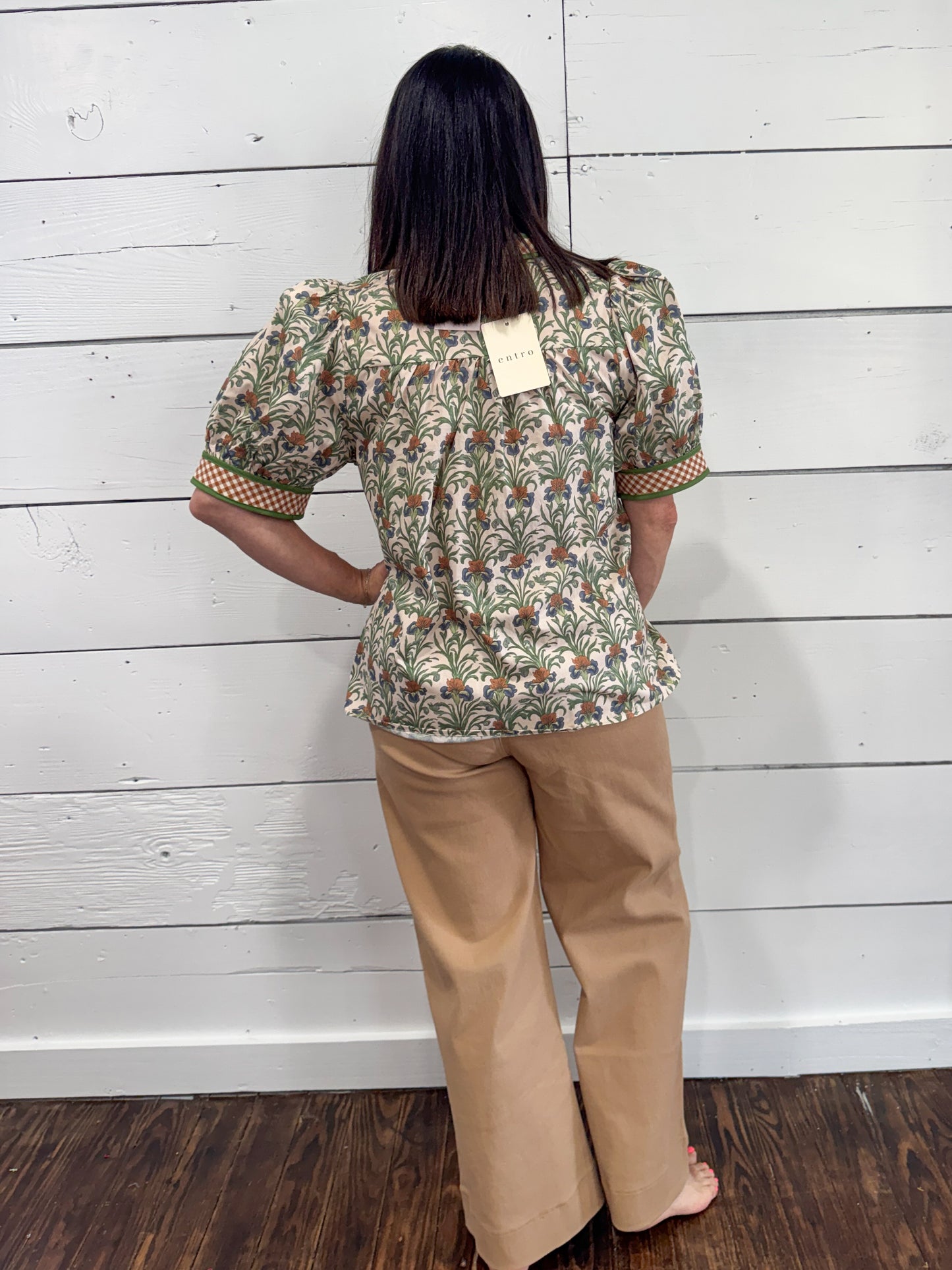 A woman stands with her back to the camera wearing a short-sleeve patterned blouse with tan wide-leg pants, posed against a white wood-paneled wall.