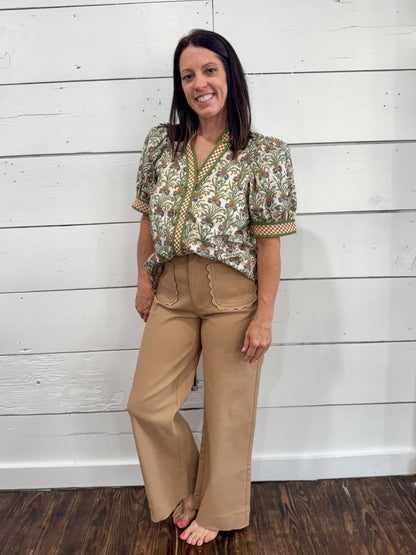 A woman stands barefoot against a white wood plank wall, wearing a short-sleeve button-front blouse with a multicolor floral and botanical print, paired with tan wide-leg pants.