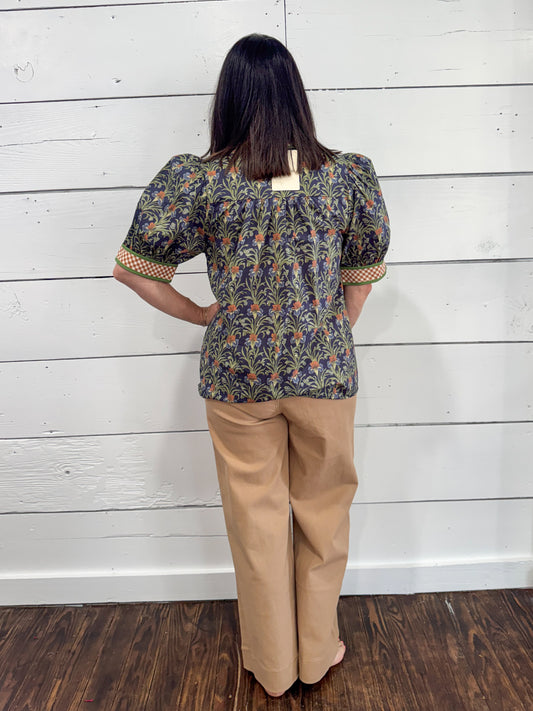 A woman stands with her back to the camera wearing a short-sleeve patterned blouse with puff sleeves and tan wide-leg pants, posed against a white wood-paneled wall on a dark wood floor.