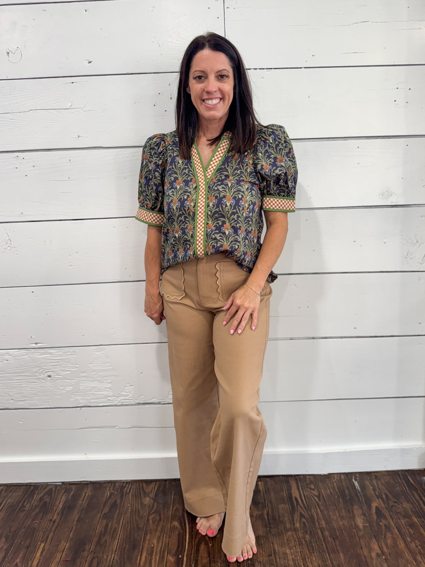 A woman stands barefoot against a white wood plank wall, wearing a short-sleeve patterned blouse with puff sleeves and tan wide-leg pants.