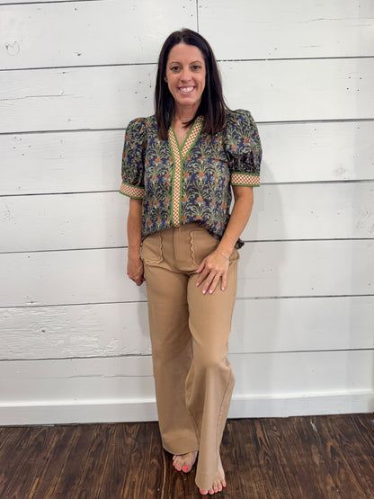 A woman stands barefoot against a white wood plank wall, wearing a short-sleeve patterned blouse with puff sleeves and tan wide-leg pants.