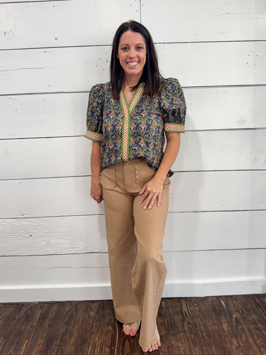 A woman stands barefoot against a white wood plank wall, wearing a short-sleeve patterned blouse with puff sleeves and tan wide-leg pants.