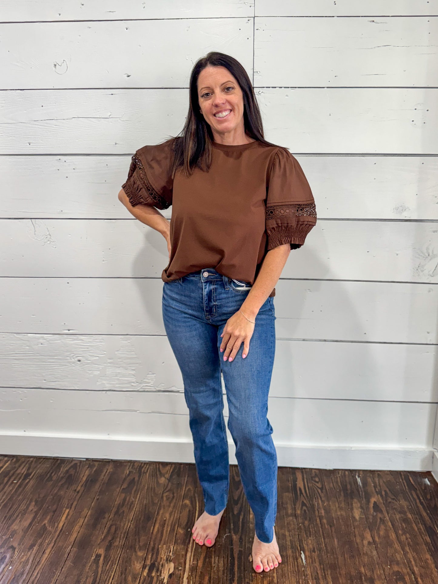 A woman modeling a brown short-sleeve top with textured puff sleeves and blue jeans, standing against a white shiplap wall on a dark wood floor.