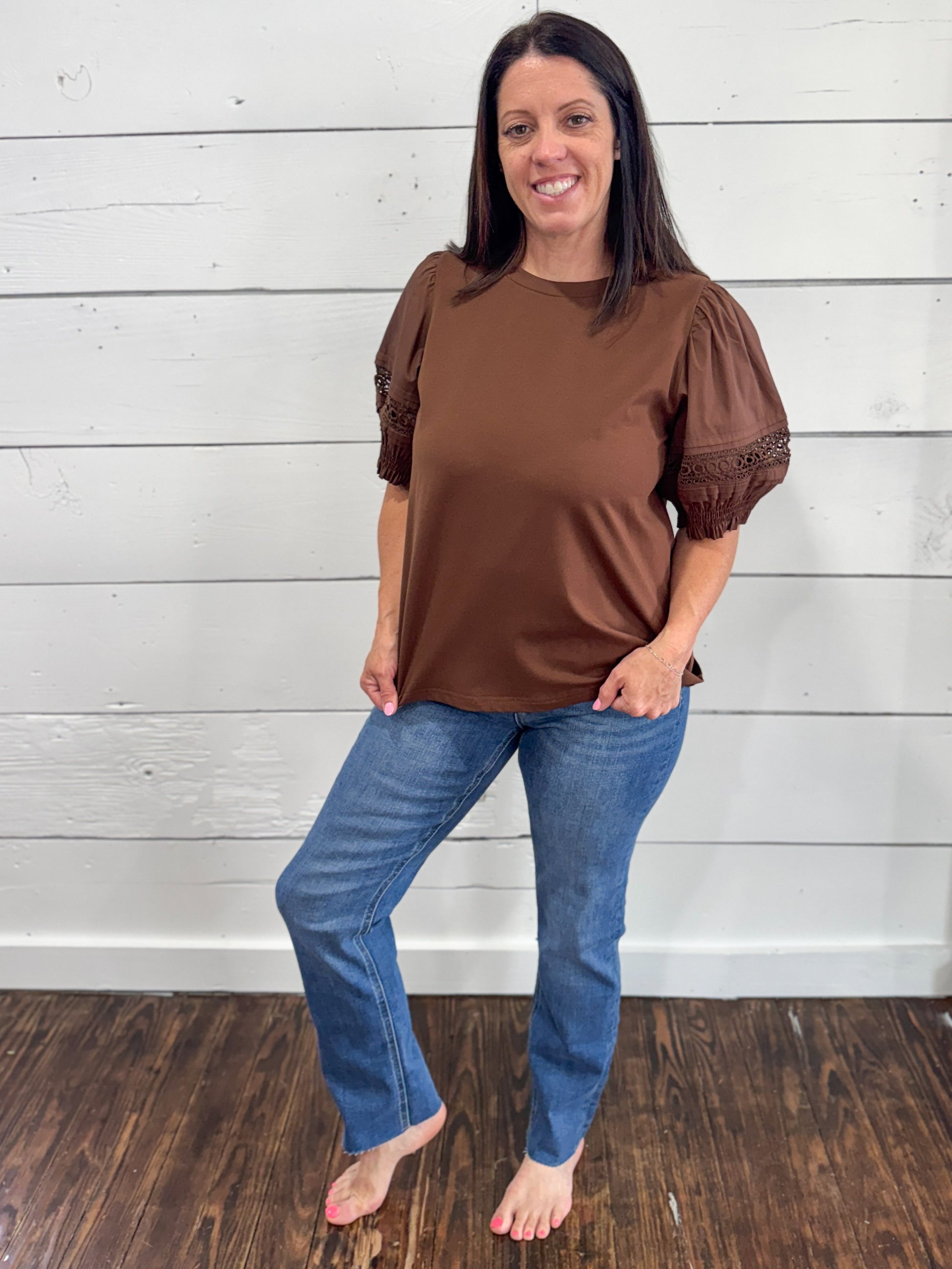 A woman modeling a brown short-sleeve top with puff sleeves and lace trim, paired with blue jeans, standing against a white wood-paneled wall on a dark wood floor.