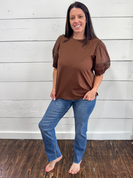 A woman modeling a brown short-sleeve top with puff sleeves and lace trim, paired with blue jeans, standing against a white wood-paneled wall on a dark wood floor.