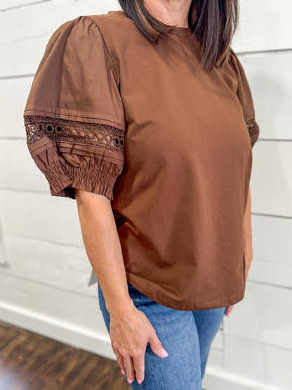 A woman wearing a brown short-sleeve blouse with puff sleeves and lace trim, paired with blue jeans, standing against a white shiplap wall.